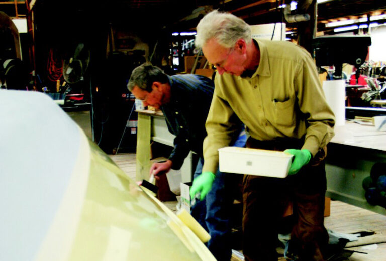 One person rolls a coat onto an overturned hull while a second follows behind with a tipping brush.
